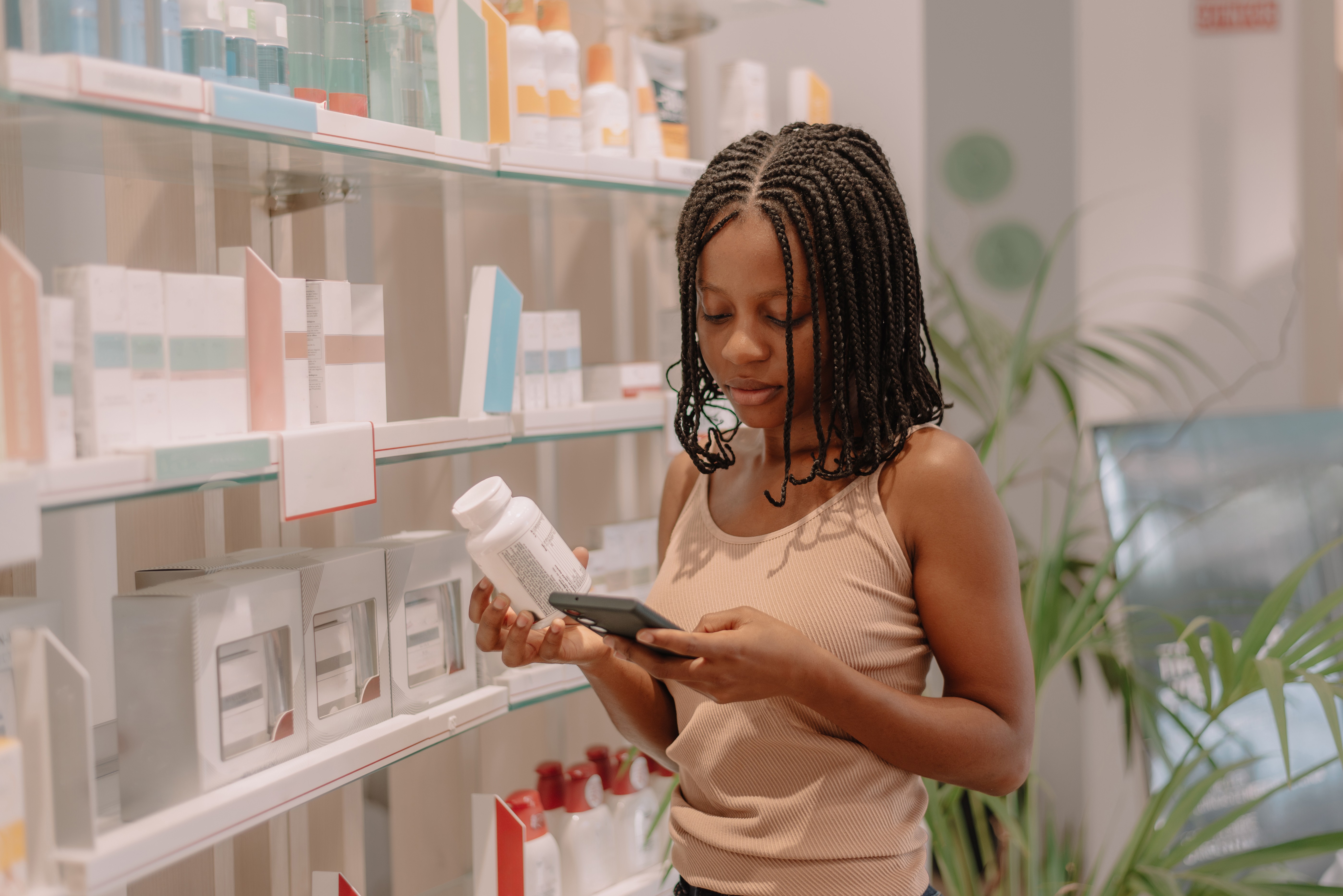 Mulher jovem com tranças, vestindo regata bege, segura um frasco branco e um celular preto, comparando informações em uma prateleira de farmácia ou loja de cosméticos