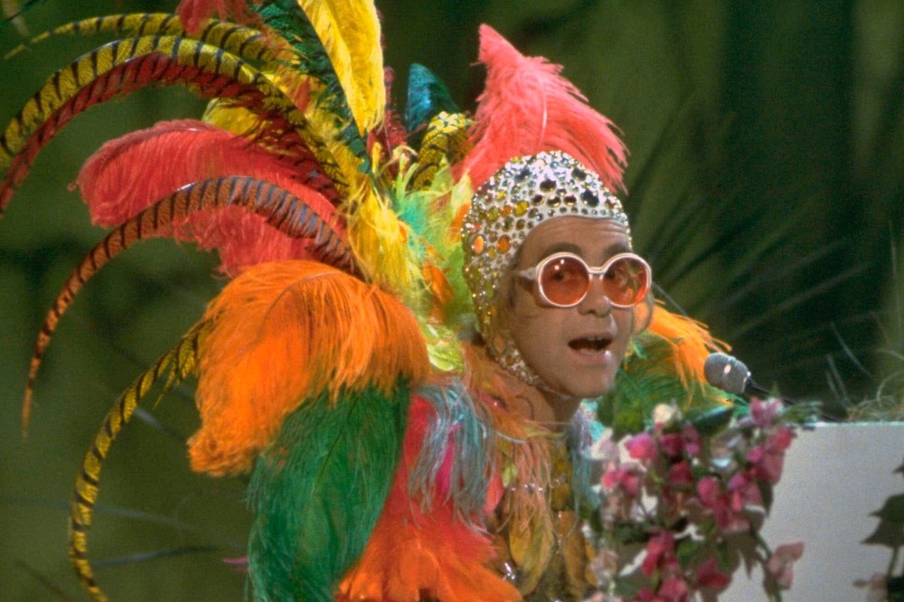 Elton John com fantasia extravagante de penas coloridas, chapéu brilhante e óculos redondos de lentes alaranjadas, cantando em um palco com flores
