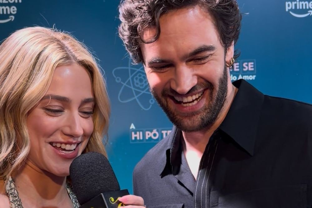 Uma mulher loira sorridente, de vestido estampado, segura um microfone preto enquanto um homem barbudo de camisa escura ri ao seu lado, em um evento com fundo azul e o logo Amazon Prime