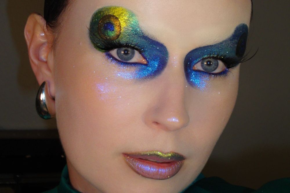 Rosto de uma mulher com maquiagem artística vibrante. Os olhos azuis têm sombras em tons de azul, verde e amarelo, com um desenho que lembra uma pena de pavão na pálpebra esquerda. Há glitter azul e roxo nas maçãs do rosto e lábios com batom metálico verde e roxo. Ela usa um brinco prateado em formato de gota na orelha esquerda