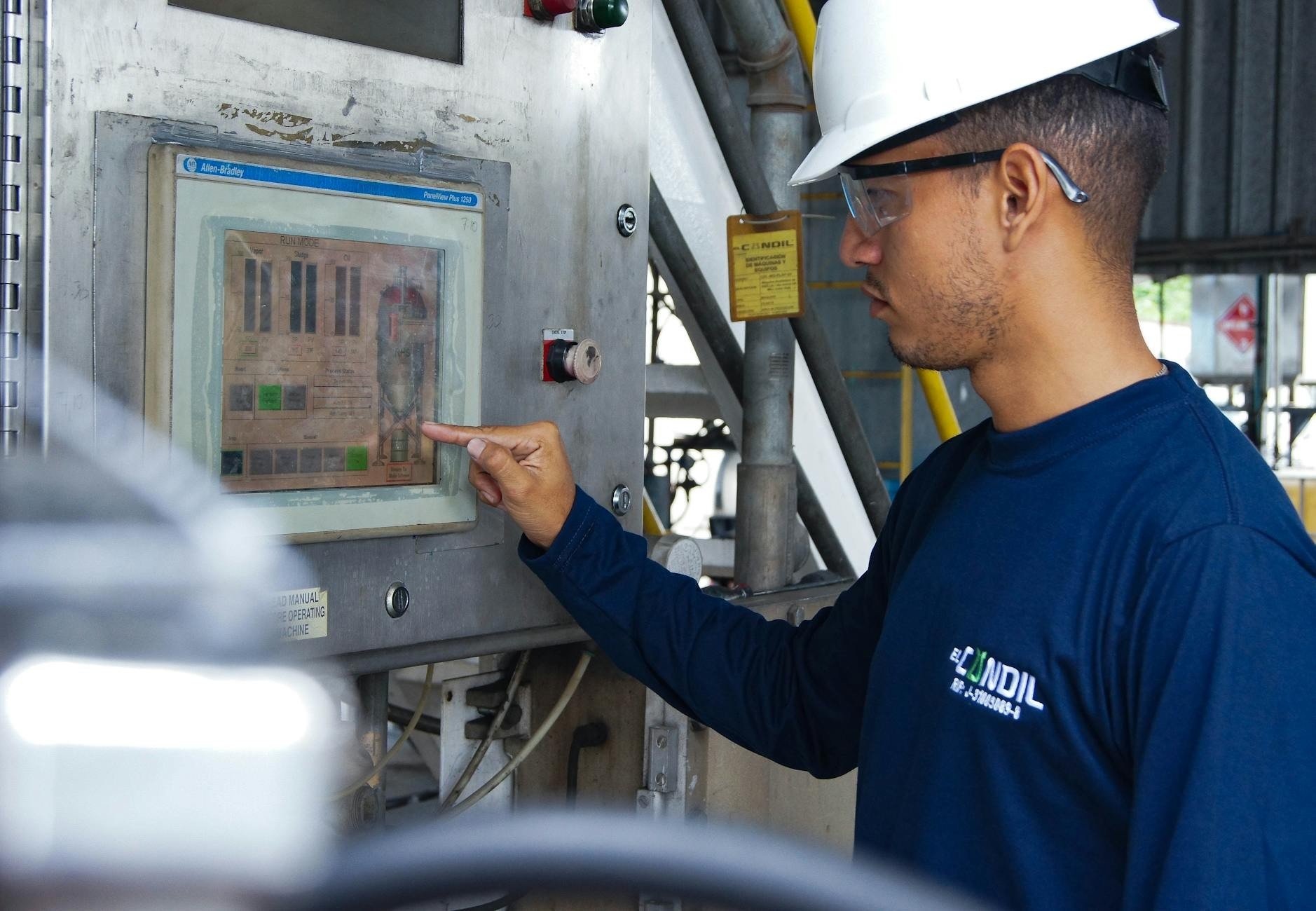 Homem jovem, de perfil, usando capacete branco e óculos de proteção, camisa azul escura, apontando para uma tela de controle industrial com gráficos e botões em um painel metálico